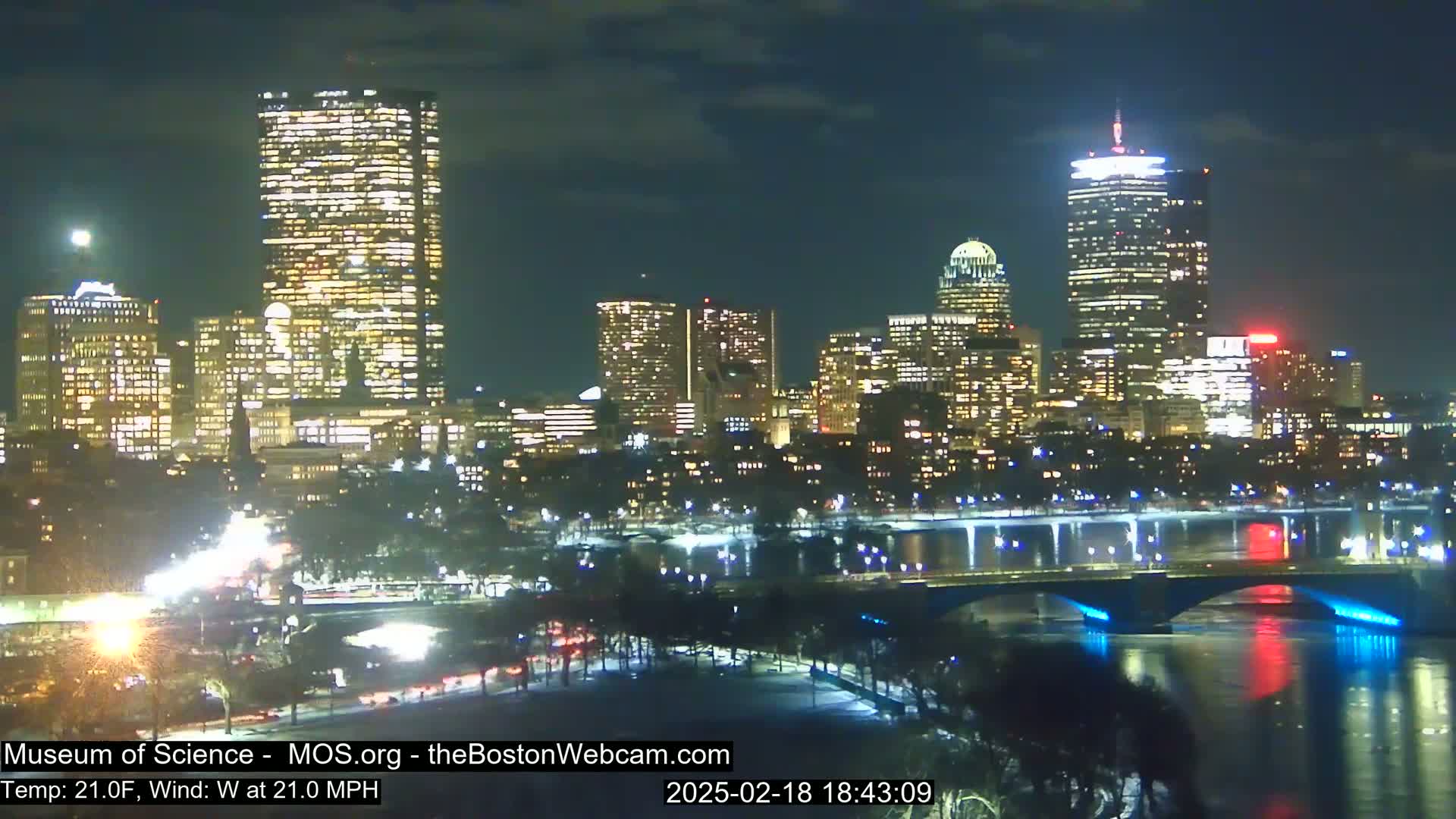 A nighttime cityscape with brightly lit skyscrapers and bridges over a river, under a clear sky with a 21°F temperature and 21 mph west wind.