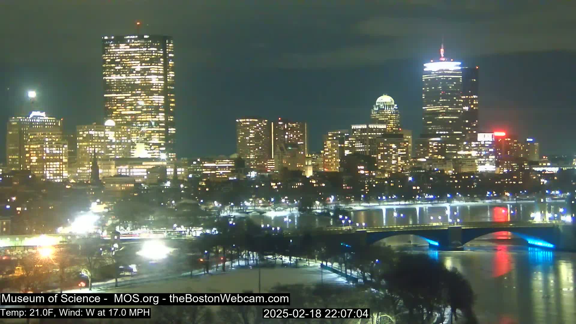 A nighttime view of a city skyline illuminated by lights, with a river reflecting the city lights and a bridge crossing it, under a clear night sky.