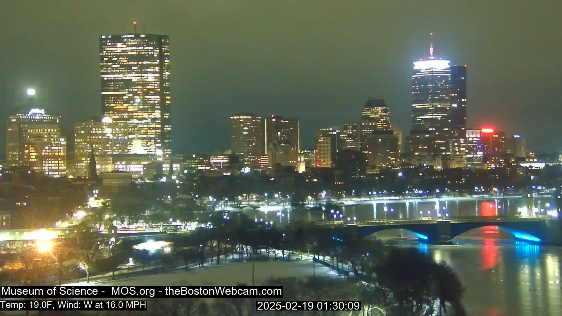 A nighttime cityscape with several illuminated skyscrapers and a bridge over a partially frozen river under a dark, overcast sky; there is snow on the ground.
