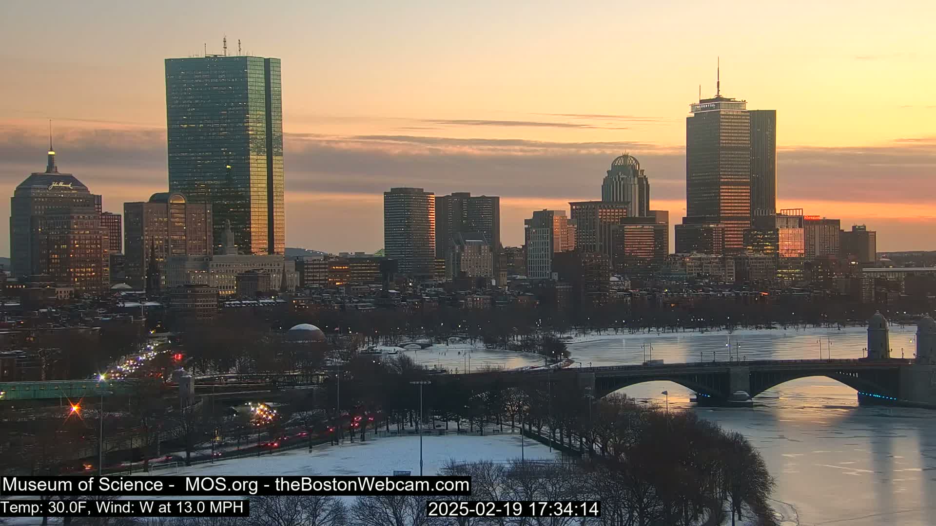 A city skyline at sunset, with snow on the ground and ice on the river, under a mostly clear sky.