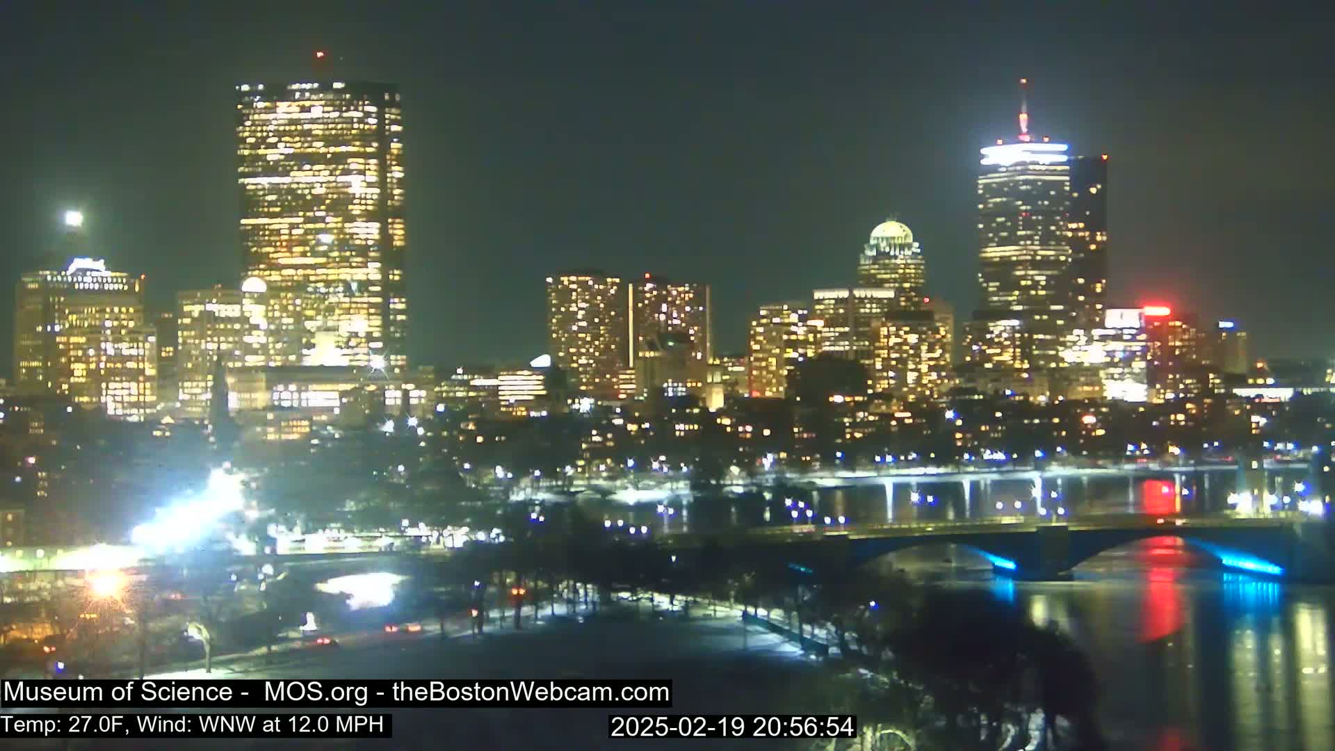 A nighttime cityscape illuminated by many lights, featuring several tall buildings across a body of water with a bridge, under a clear night sky.