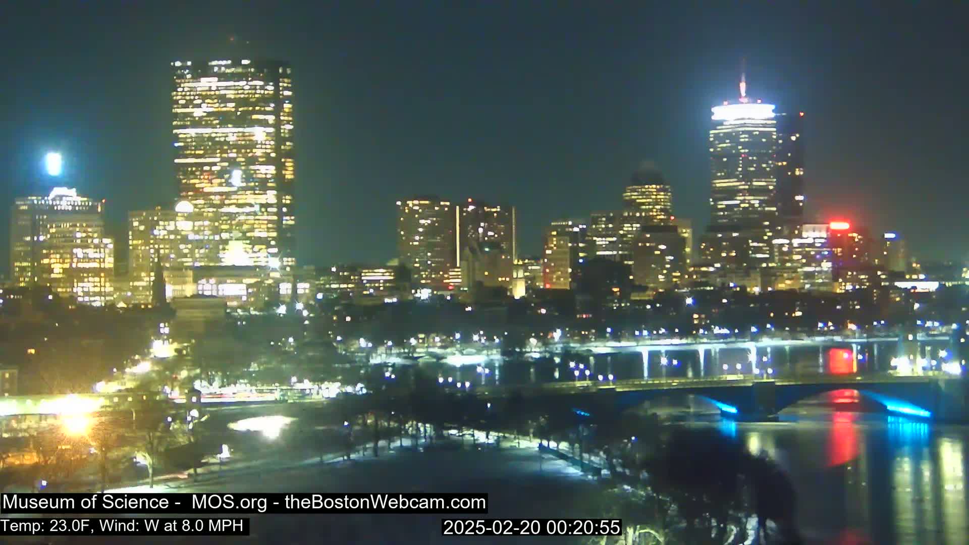 A nighttime view of a city skyline across a river, with a bridge illuminated in blue and red lights, under a clear sky, and a temperature of 23 degrees Fahrenheit and a west wind of 8 mph.