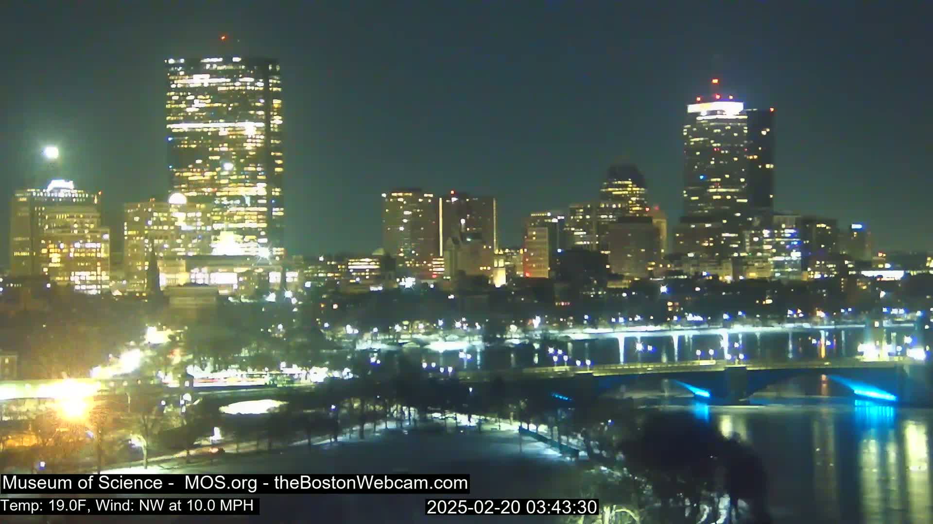 A nighttime cityscape with illuminated skyscrapers and bridges over a dark river, under a clear sky with a visible moon and a northwest wind at 10 mph and a temperature of 19°F.