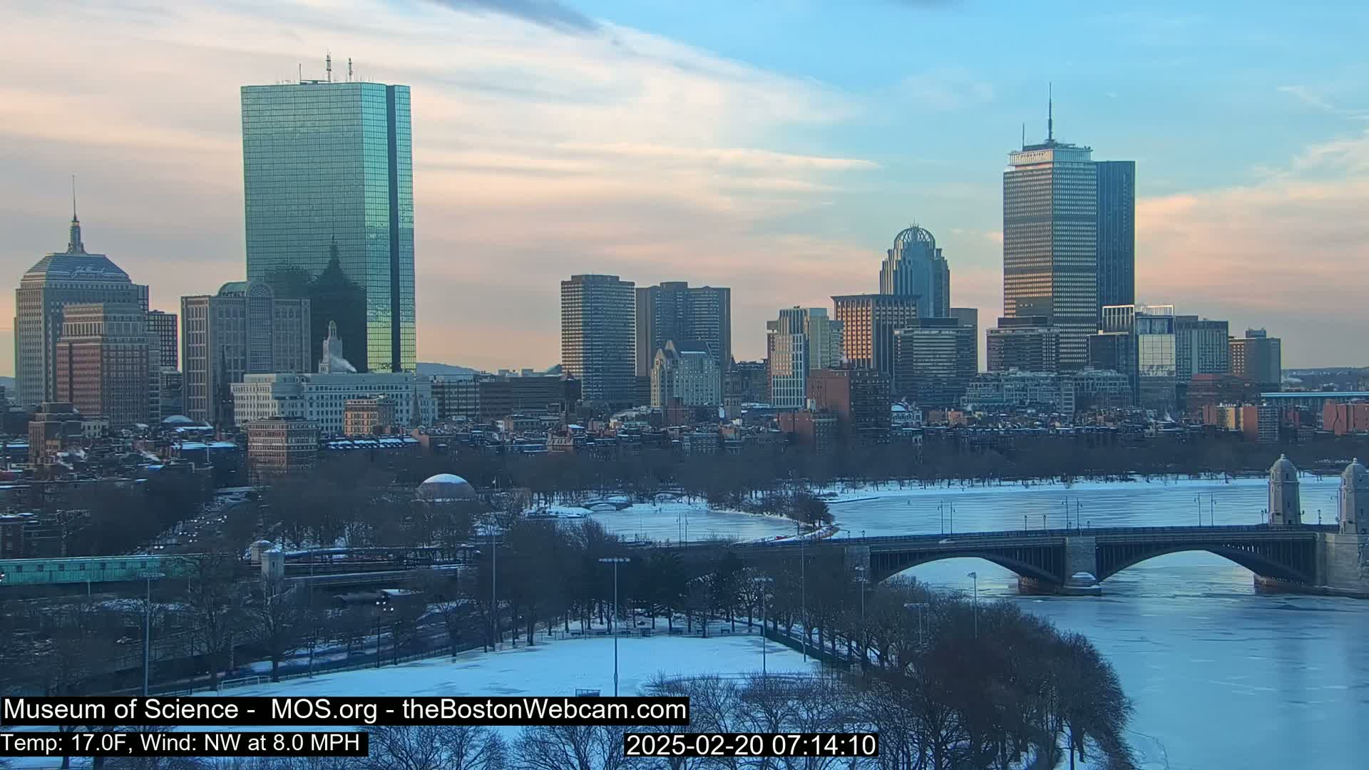 A snow-covered Boston cityscape at sunset, with a partially frozen river and a bridge in the foreground.