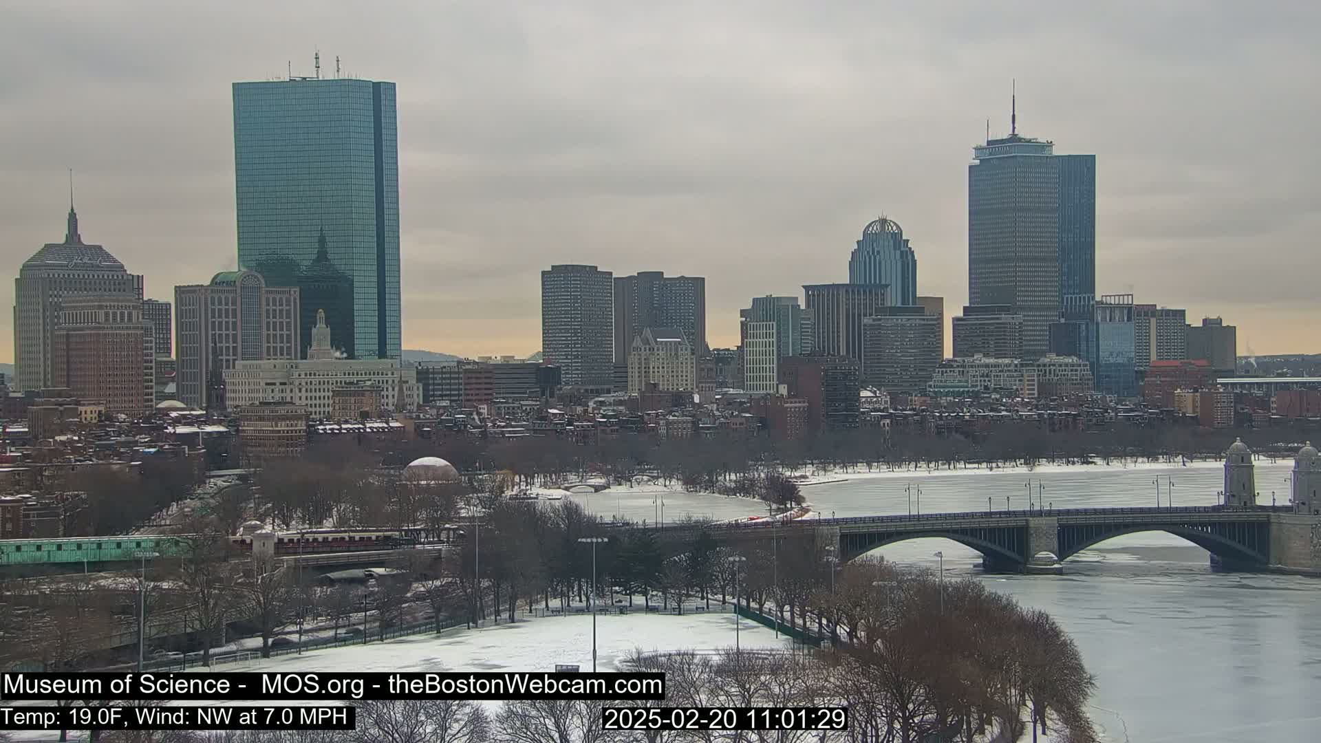 A snow-covered city skyline with a frozen river and bridge under a cloudy sky.