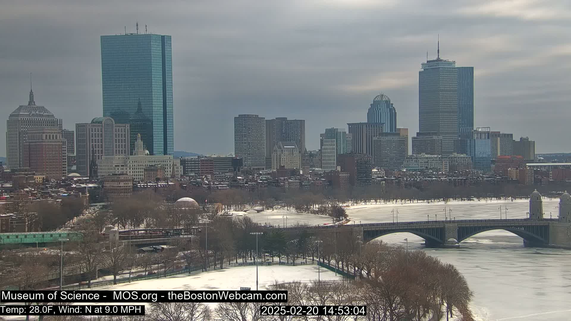 A snow-covered cityscape with a frozen river and a bridge, under an overcast sky.