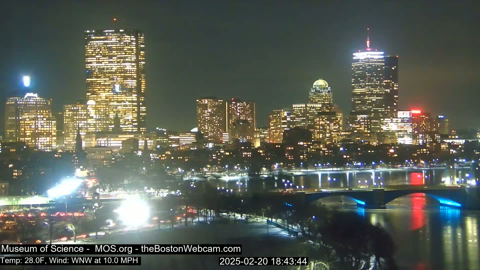 A nighttime cityscape with brightly lit skyscrapers and a bridge reflected in a dark river under a clear sky.