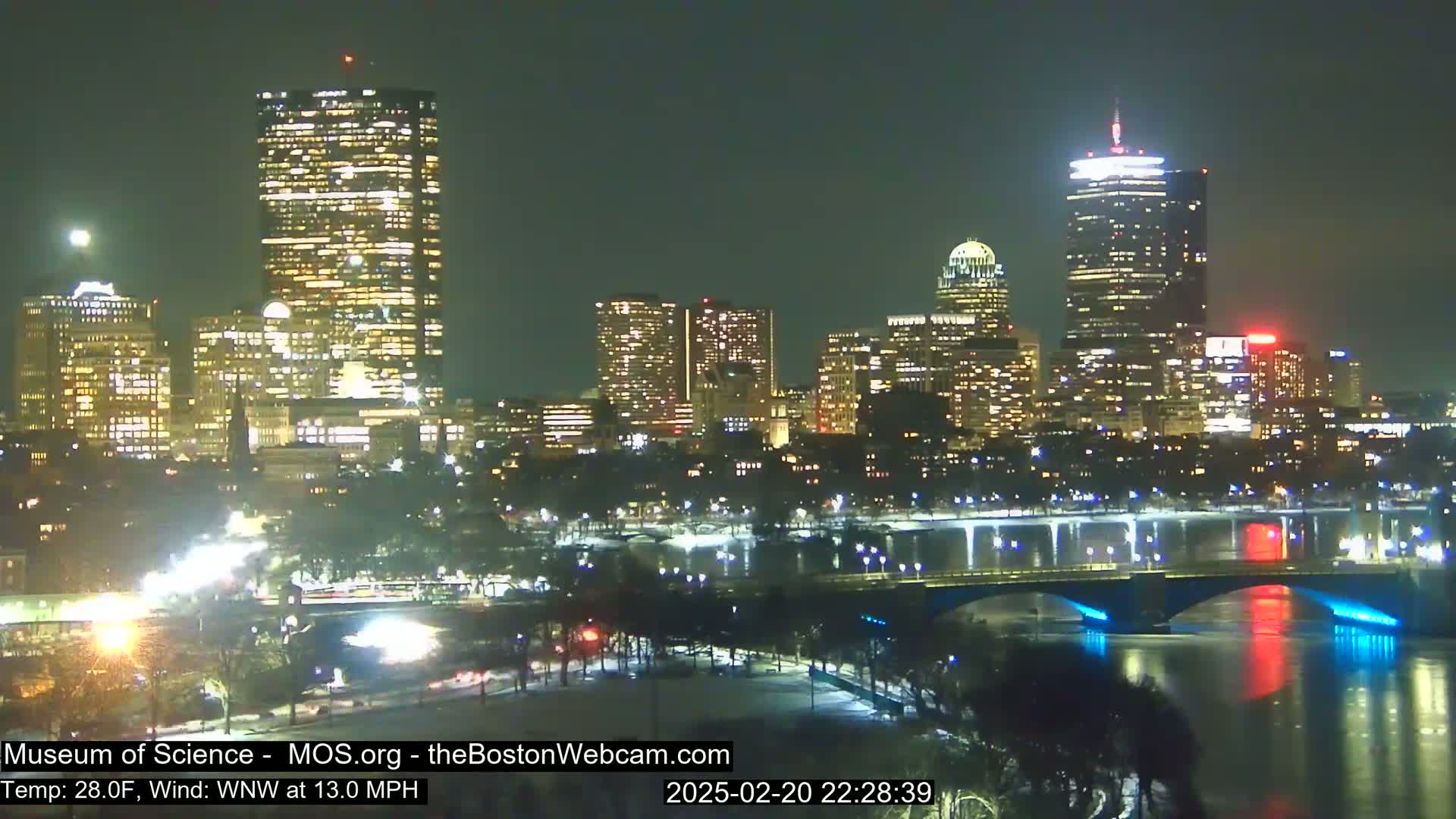 A nighttime view of a city skyline illuminated by lights, with a bridge spanning a dark river under a clear sky, and a temperature of 28°F and a wind of 13 mph from the west-northwest.