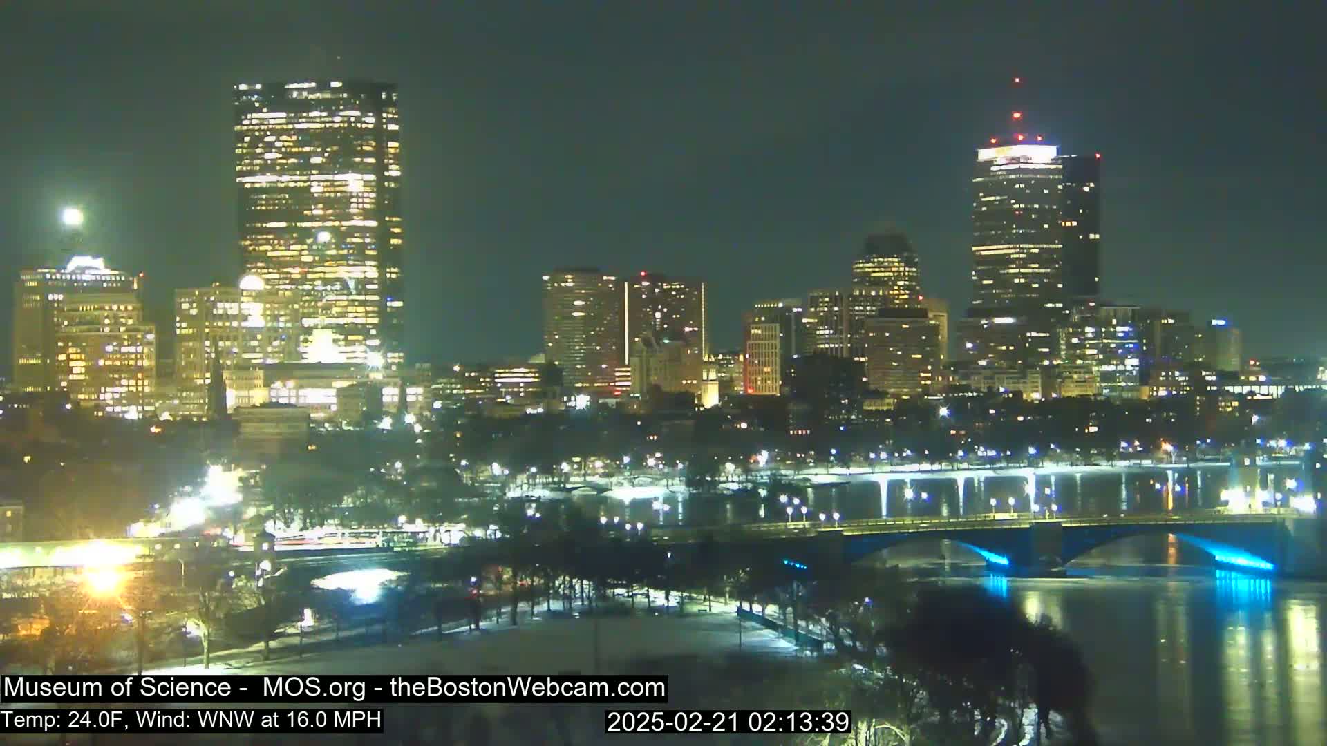 A nighttime view of a city skyline across a partially frozen river, under a clear sky with a west-northwest wind at 16 mph and a temperature of 24 degrees Fahrenheit.