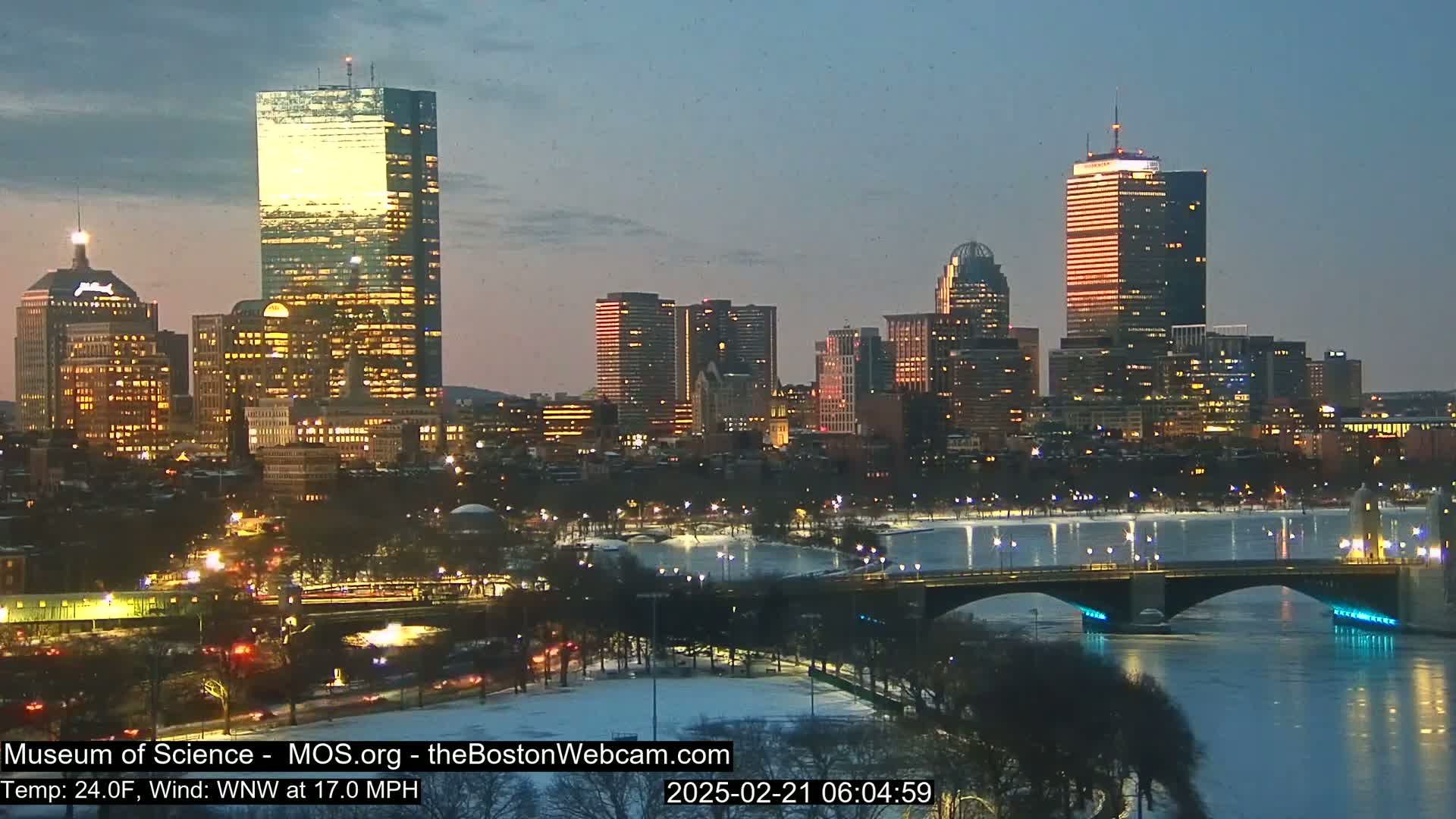 A nighttime view of a city skyline with illuminated buildings, a bridge over a partially frozen river, and trees dusted with snow under a twilight sky with a wind of 17 mph.