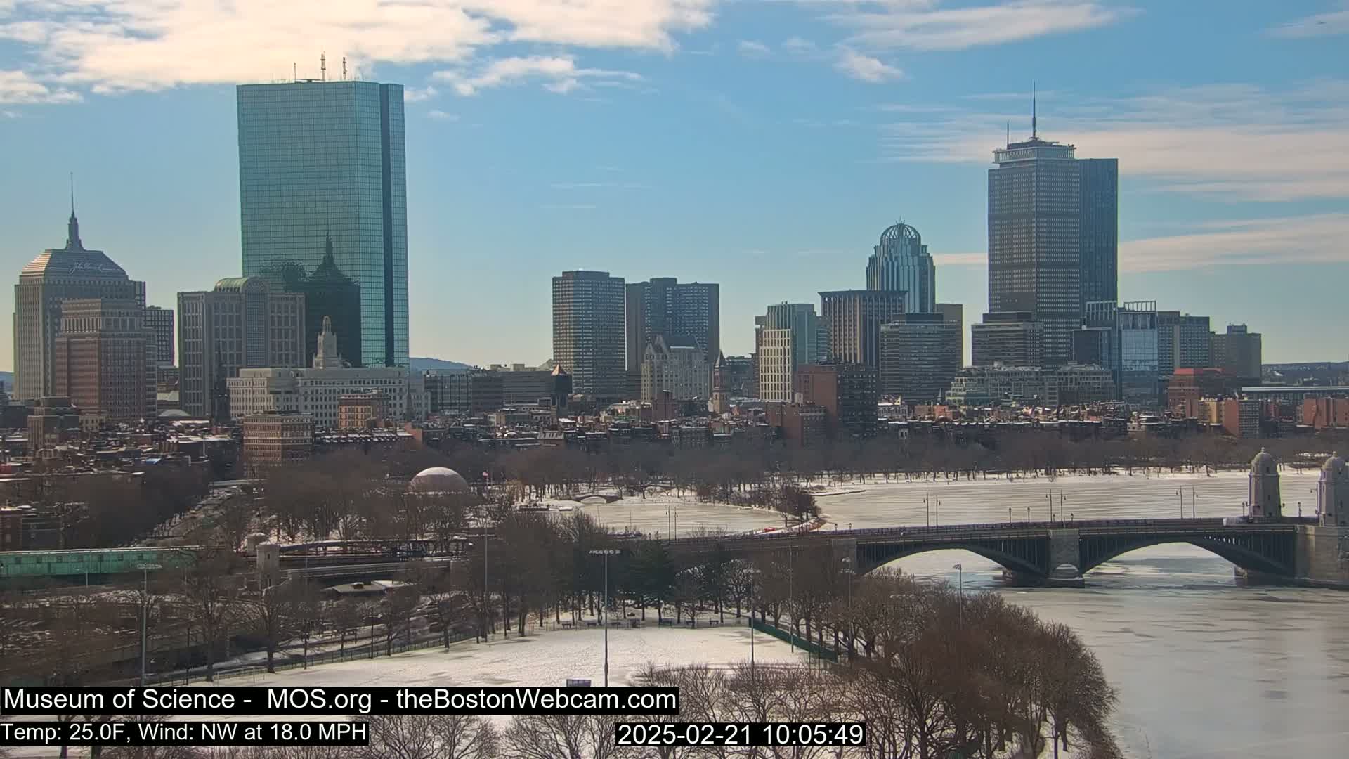 A snow-covered cityscape with a river partially frozen over, under a partly cloudy sky.
