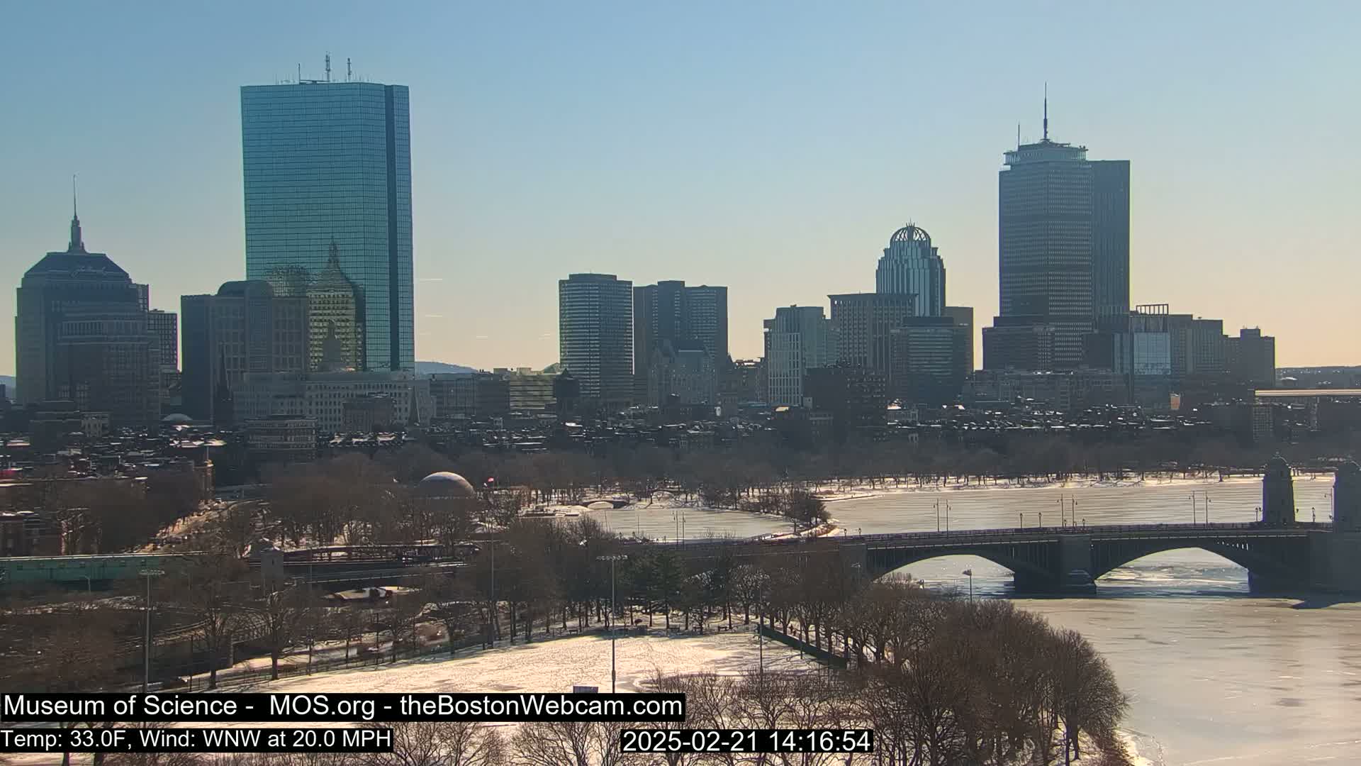 A sunny cityscape with snow-covered ground and a partially frozen river, featuring a bridge and numerous skyscrapers under a clear sky.