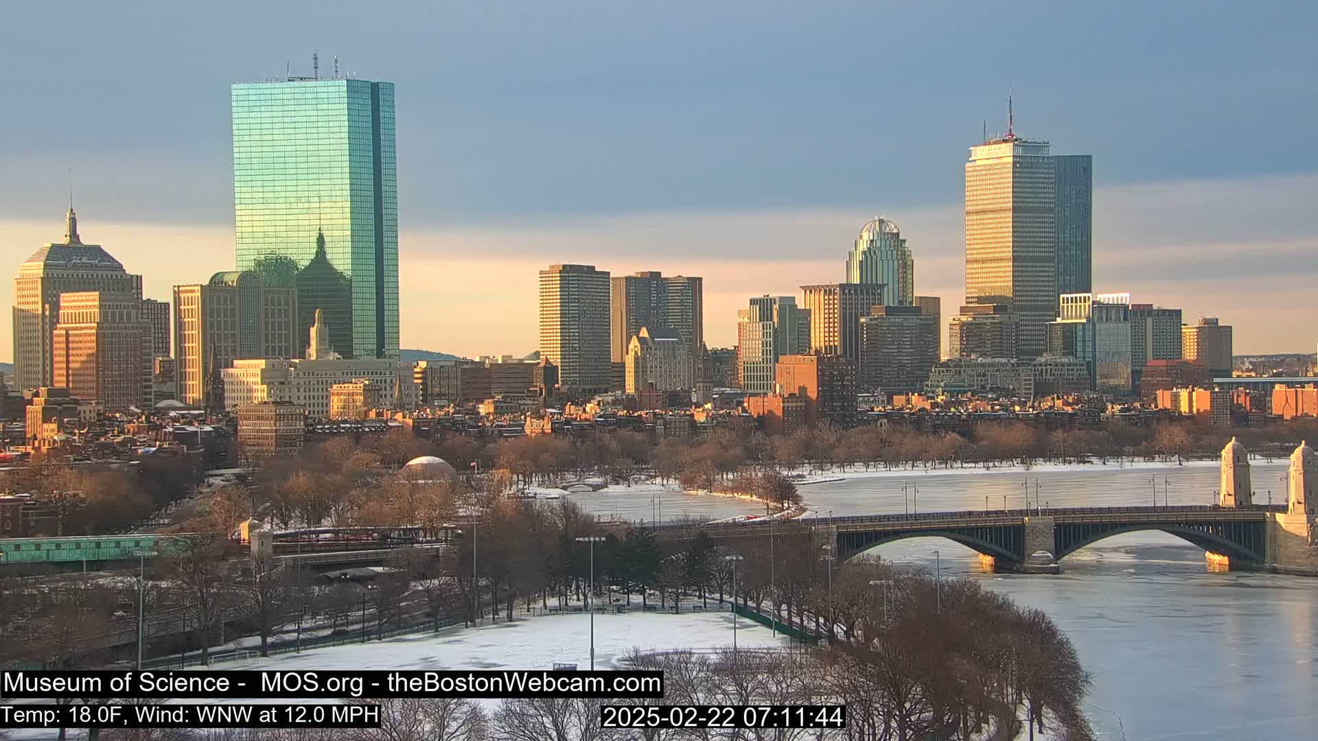 A snow-covered city skyline with a bridge spanning a partially frozen river under a partly cloudy, sunrise sky.