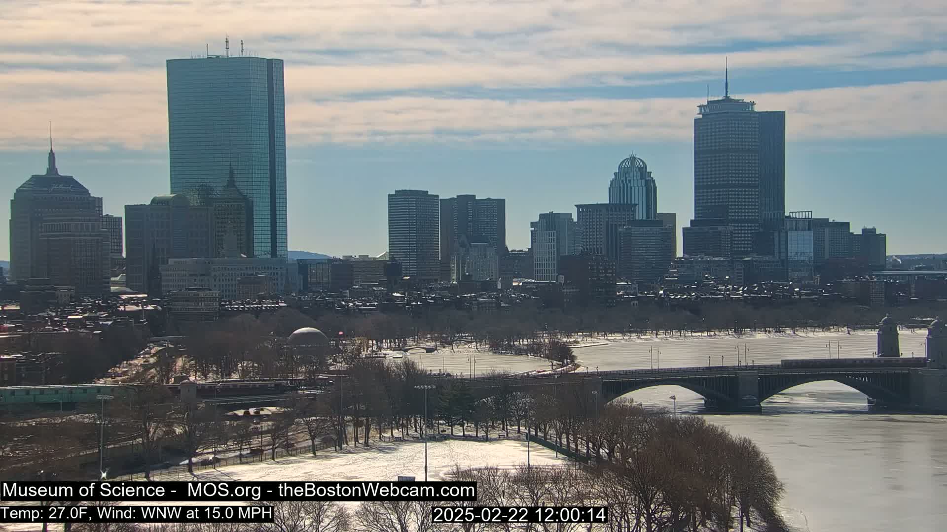 A snow-covered cityscape with a partially frozen river and a bridge, under a partly cloudy sky.