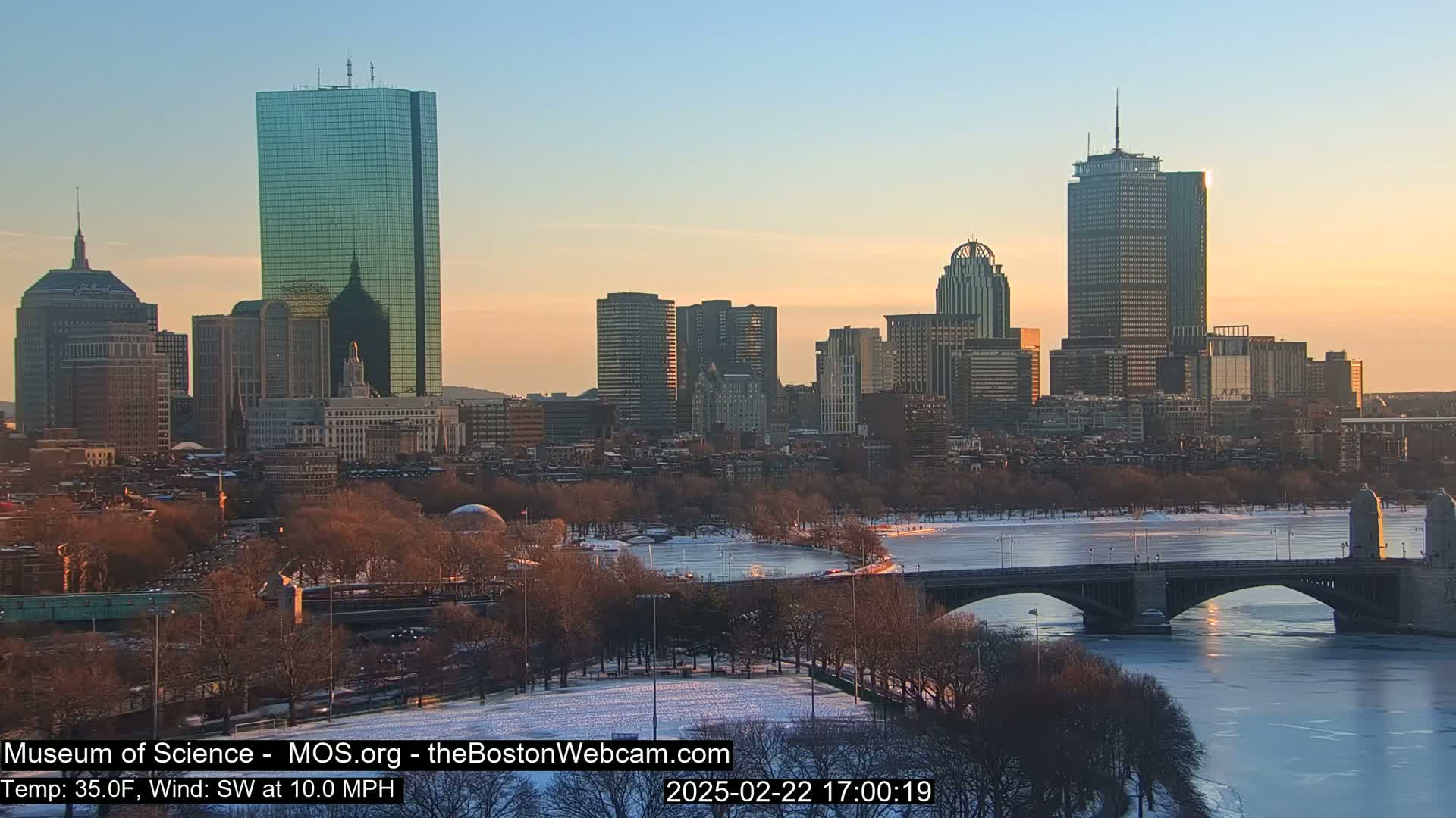 A snow-covered city skyline at sunset, with a partly frozen river and bridge in the foreground, under a clear sky.