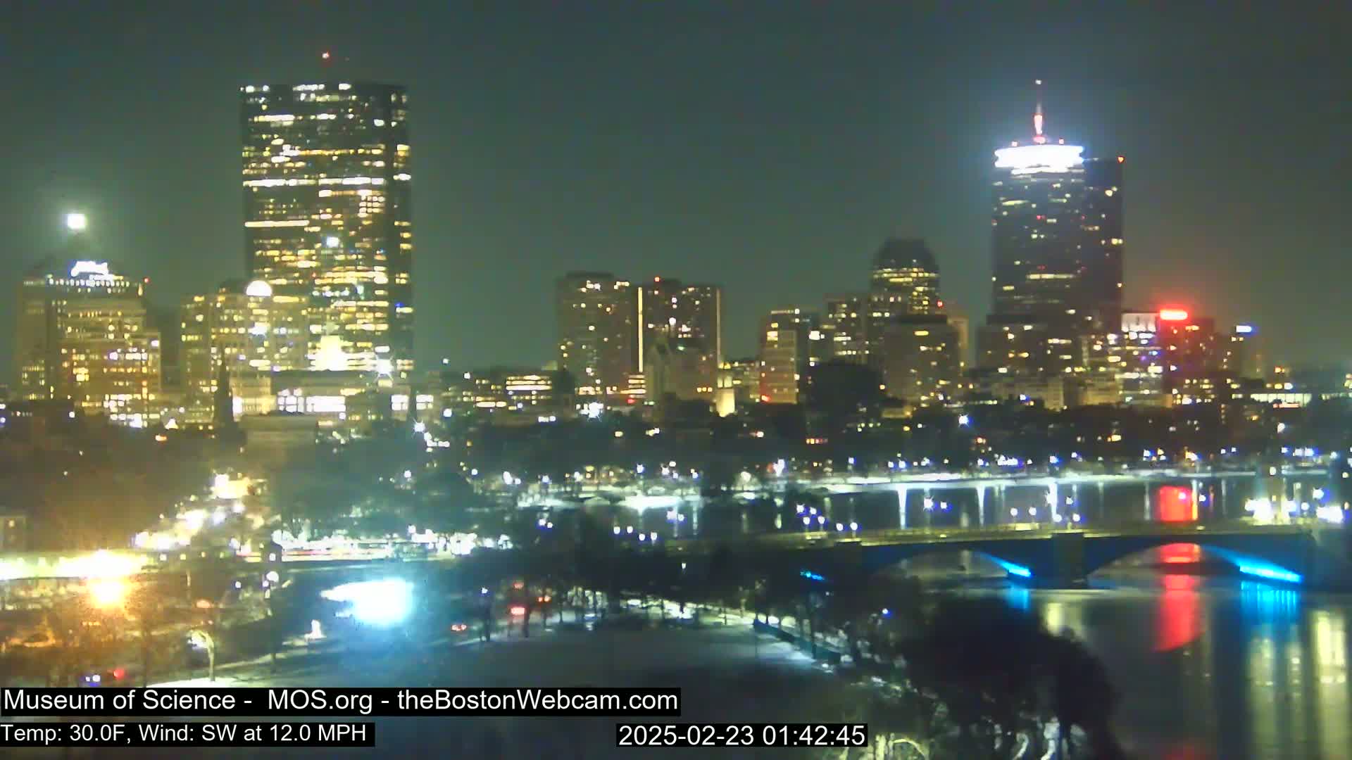 A nighttime view of a city skyline across a river, with a bridge illuminated in blue and red lights, under a clear, cool night sky.