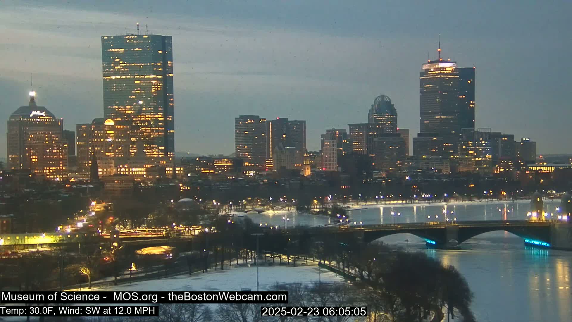 A nighttime cityscape with illuminated buildings and a bridge spanning a partially frozen river under a twilight sky.