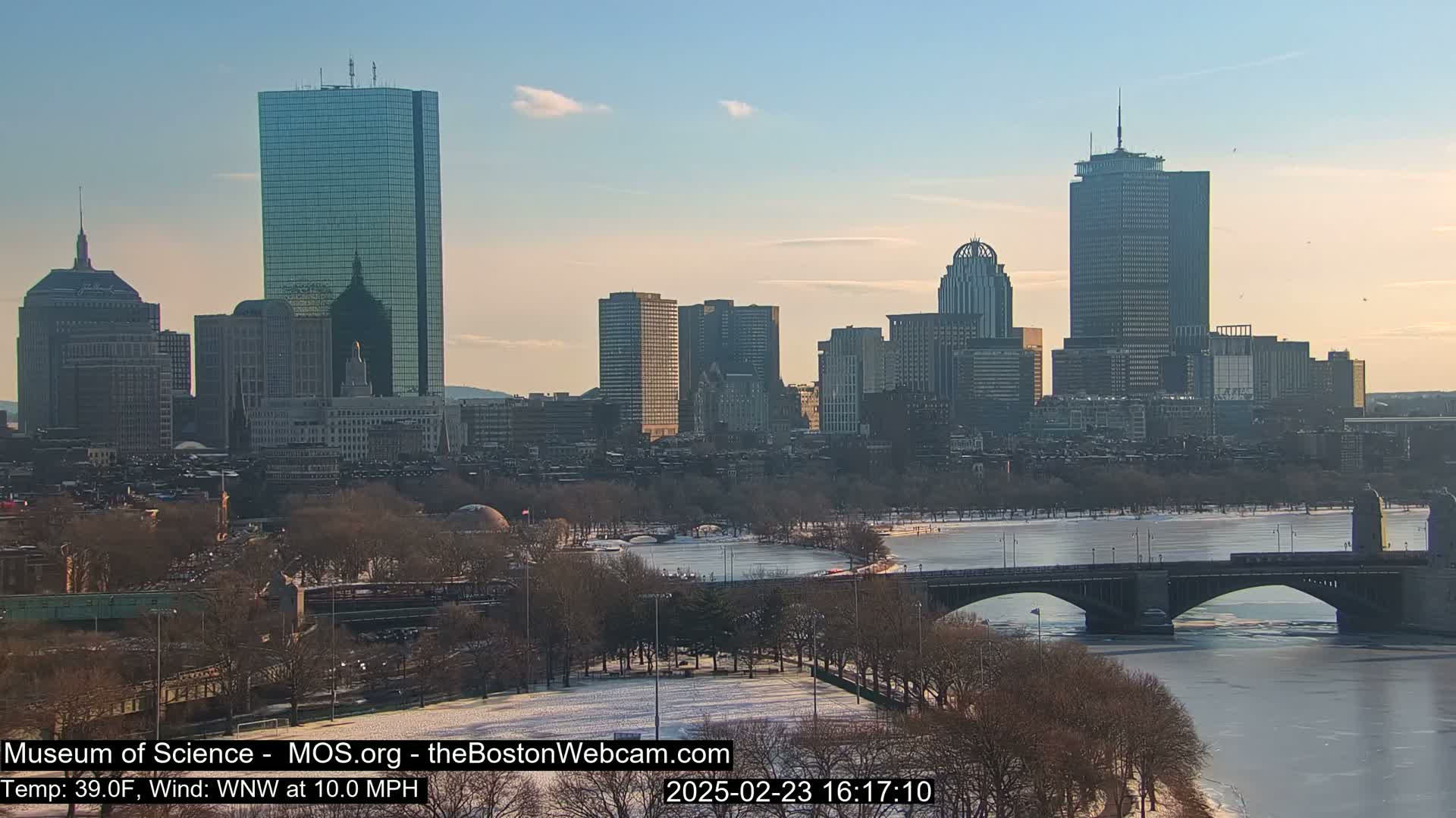 A partly frozen river flows beneath a bridge in front of a city skyline under a clear, twilight sky.