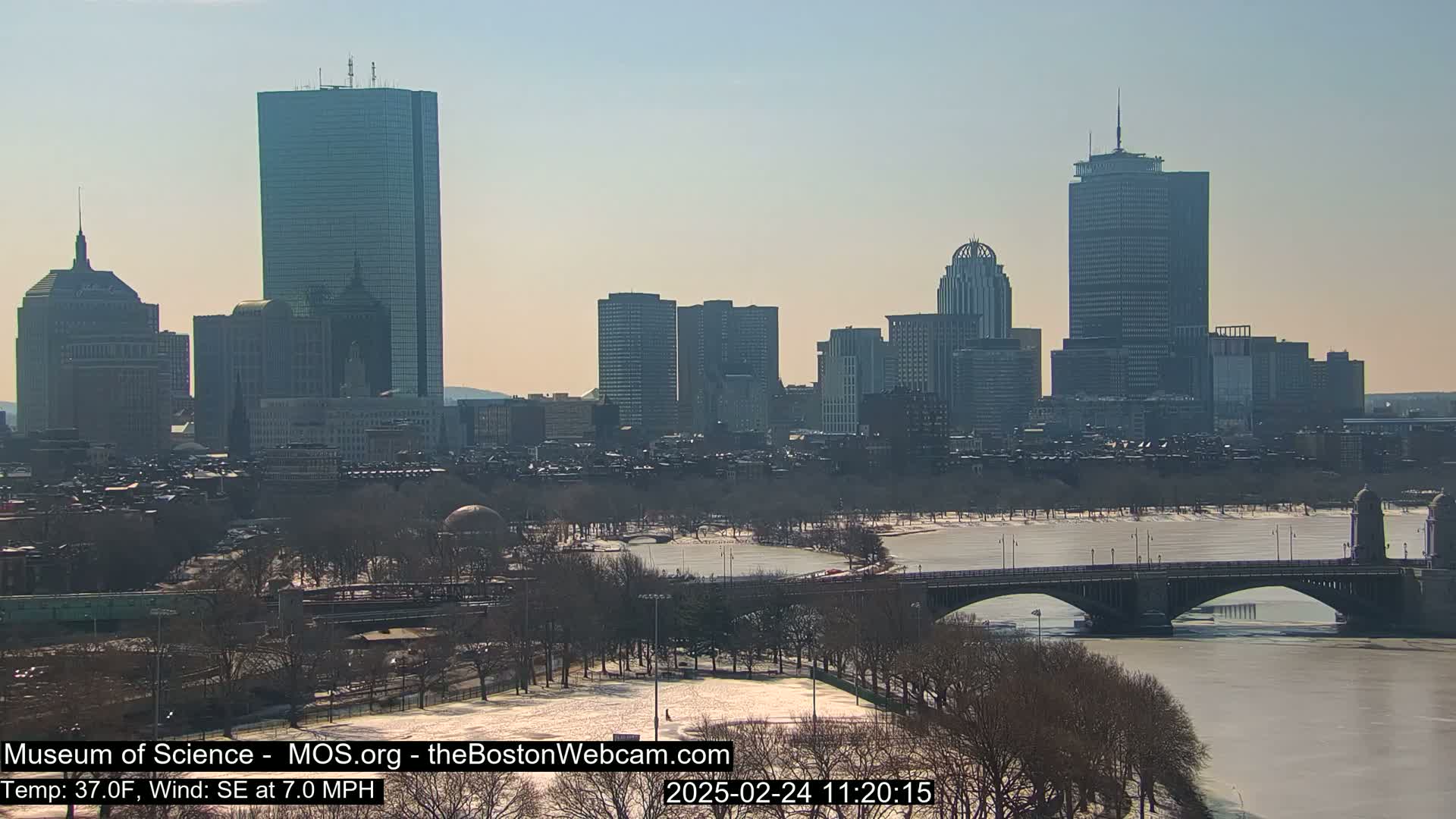 A sunny day in a city with snow-covered ground and a partially frozen river, showcasing a skyline of tall buildings and a bridge.