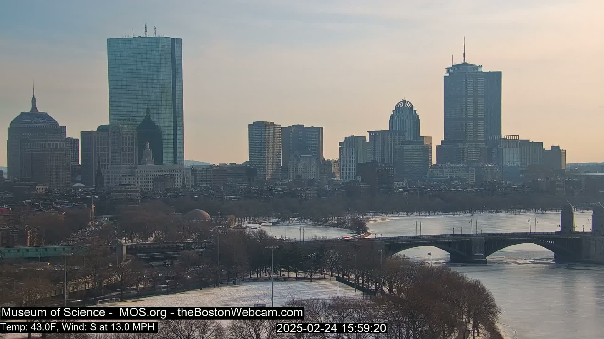 A cityscape with tall buildings, a partially frozen river, and a bridge under a partly cloudy sky.