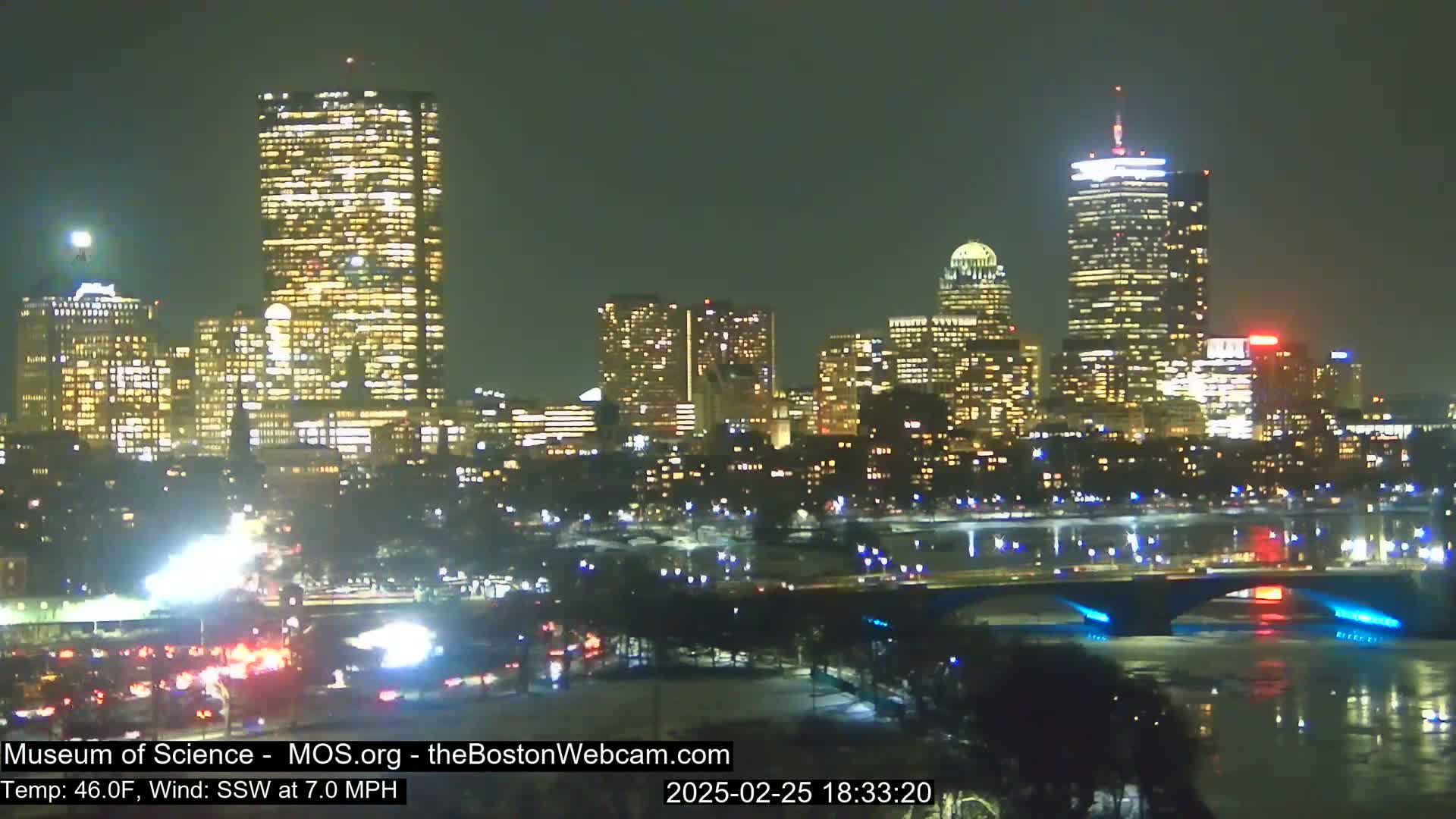 A brightly lit cityscape at night, viewed from across a river with a bridge, under a clear sky with a temperature of 46 degrees Fahrenheit and a south-southwesterly wind.
