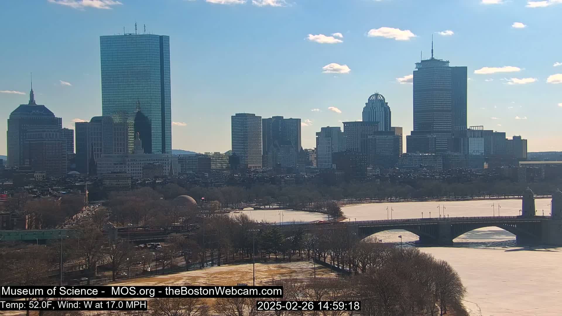 A sunny cityscape with a partially frozen river and a bridge under a mostly clear sky.