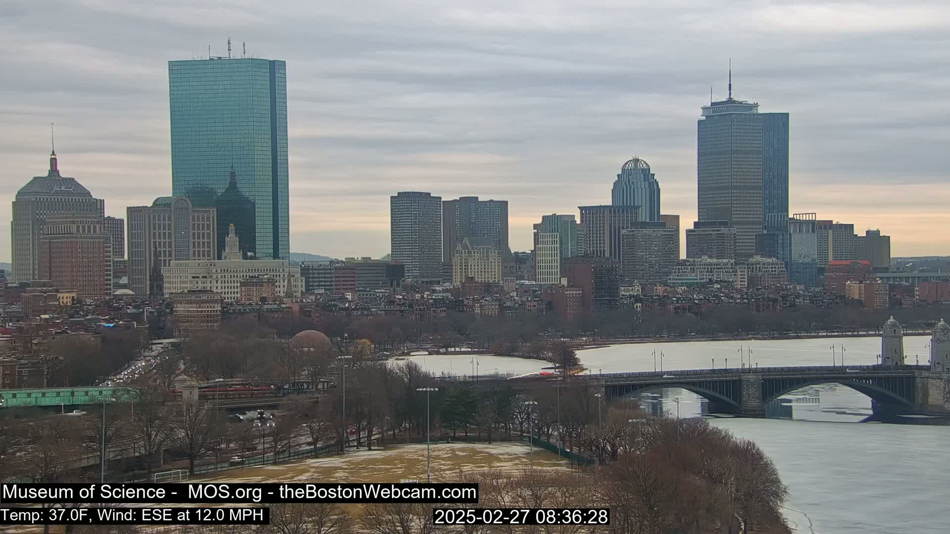 A city skyline with numerous skyscrapers and buildings is seen across a partially frozen river with a bridge, under an overcast sky.