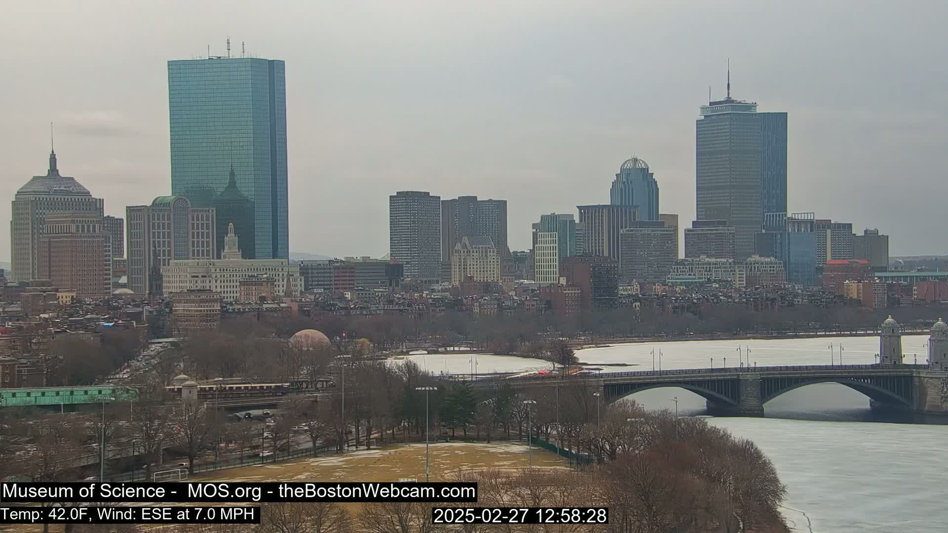 A city skyline with numerous skyscrapers is visible across a partially frozen river, under an overcast sky.