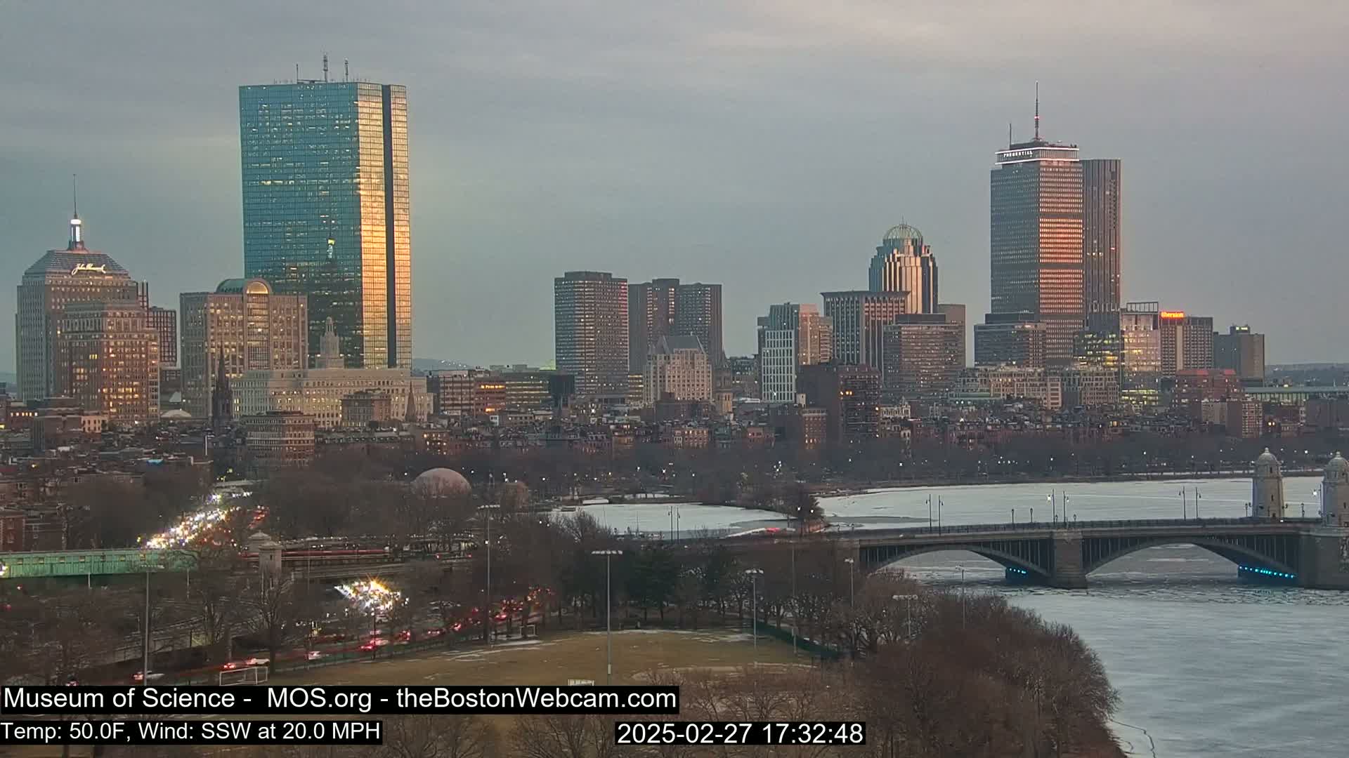 A cityscape at sunset, with a partially frozen river and bridge in the foreground, under a mostly cloudy sky and a 20 mph wind.