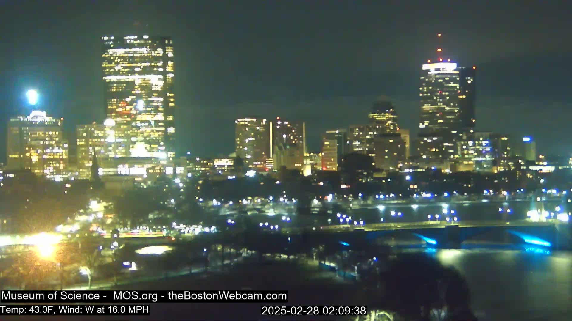 A nighttime view of a city skyline across a body of water, under a clear sky with a 16 mph west wind and a temperature of 43°F.