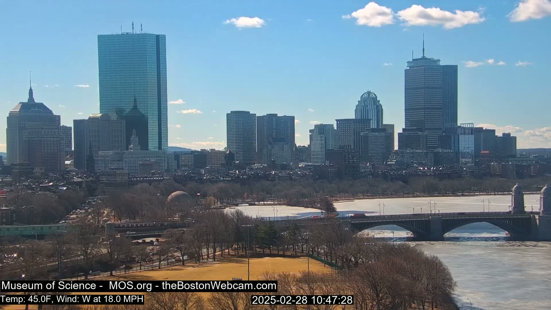 A sunny cityscape with several skyscrapers is seen across a partially frozen river, with a bridge in the foreground and bare trees lining the riverbanks.