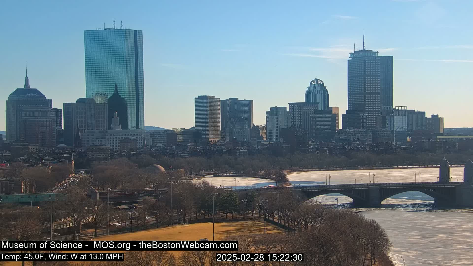 A city skyline with several skyscrapers is visible across a partly frozen river, under a clear blue sky.