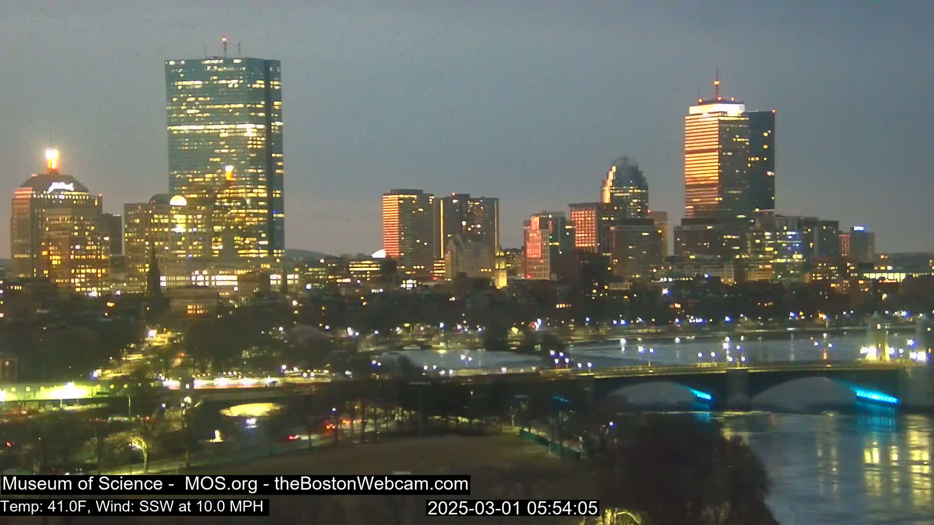 A nighttime cityscape with illuminated skyscrapers and bridges over a dark river under a dusky sky, with a temperature of 41°F and a south-southwesterly wind of 10 mph.