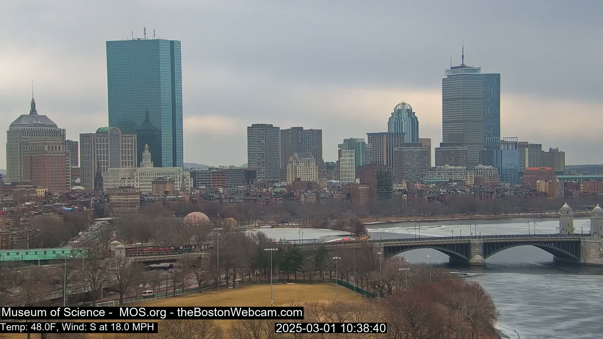 A city skyline with several tall buildings is seen across a river partially covered in ice under an overcast sky.
