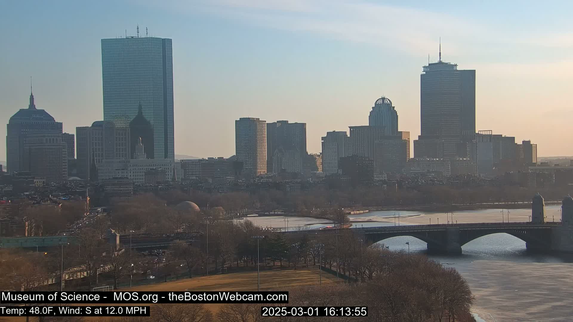 A city skyline with numerous skyscrapers and a bridge over a partially frozen river under a clear, dusky sky.