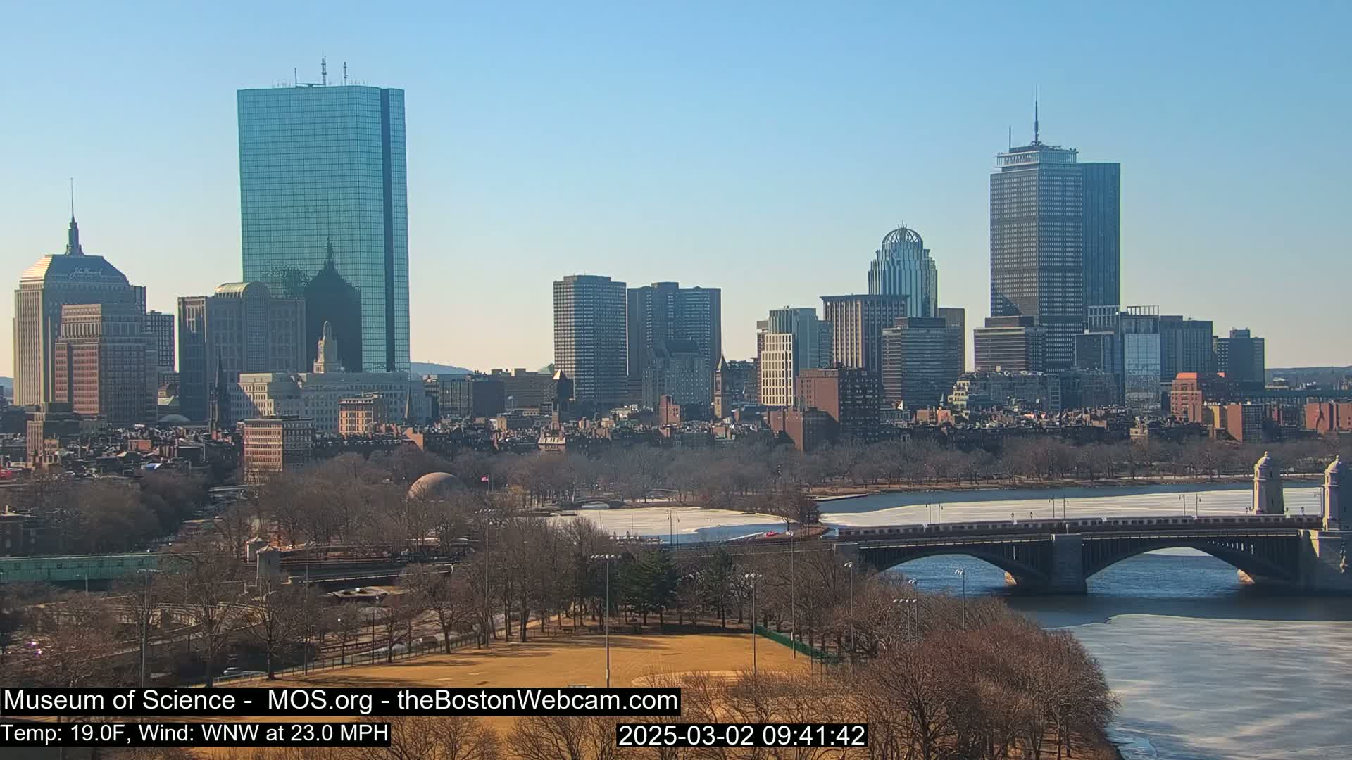 A sunny cityscape with a river and bridge, partially frozen, under a clear blue sky and a strong wind.