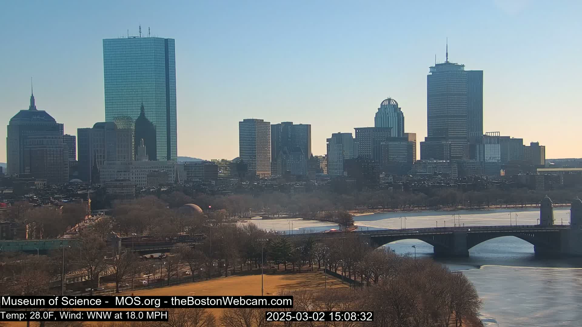 A city skyline with numerous skyscrapers is seen across a partially frozen river and bridge under a clear, sunny sky.