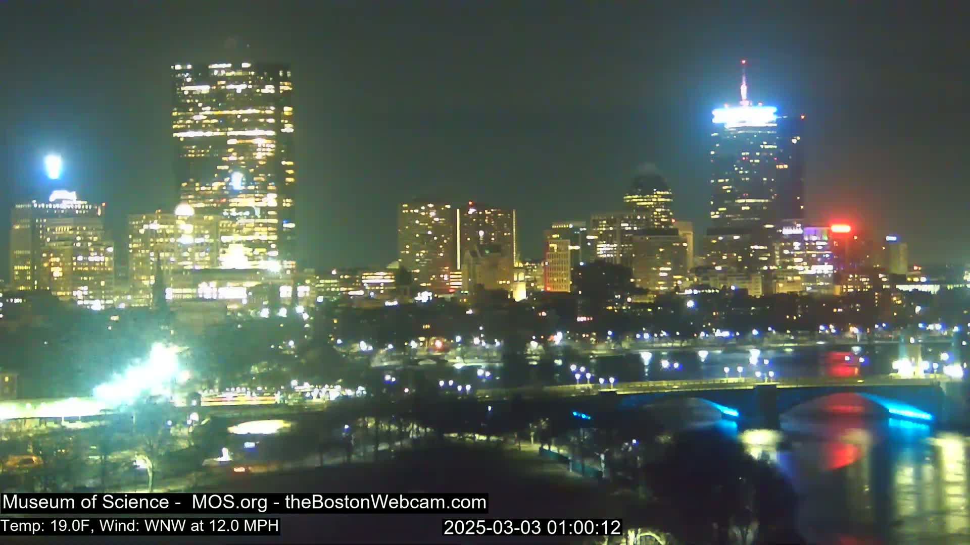 A nighttime cityscape with illuminated buildings and a bridge over a body of water, under a clear sky with a 19°F temperature and a 12 mph west-northwest wind.