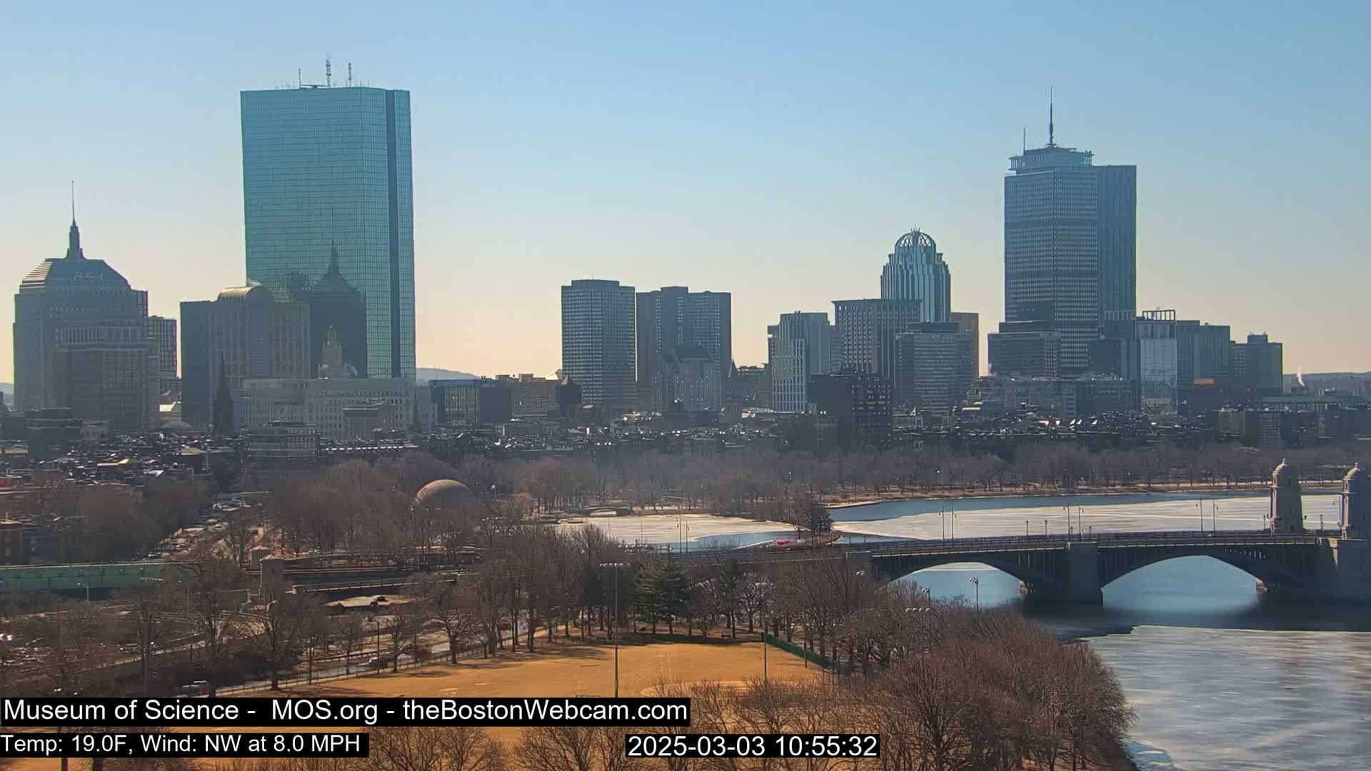 A sunny cityscape with a river partially frozen over, a bridge, and bare trees under a clear sky.