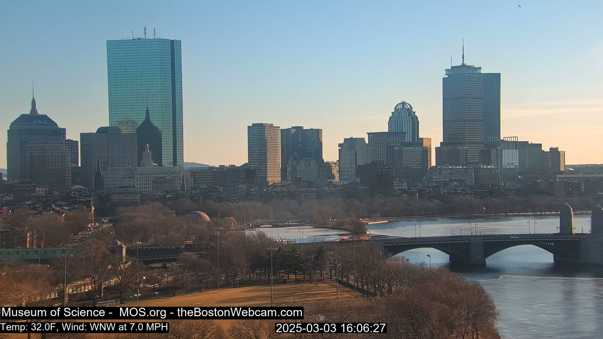 A clear, cold day reveals a cityscape with several skyscrapers and a bridge spanning a partially frozen river, with bare trees lining the riverbanks and a park visible in the foreground.