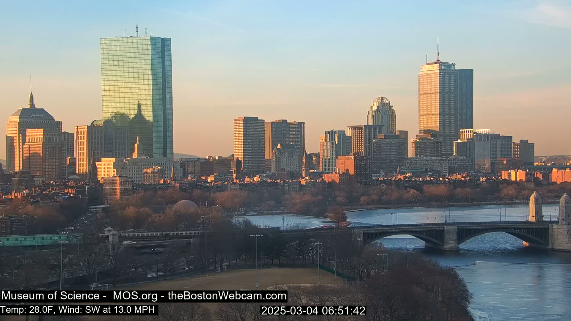A city skyline at sunrise, with a river, bridge, and some partially frozen water visible under a clear sky.
