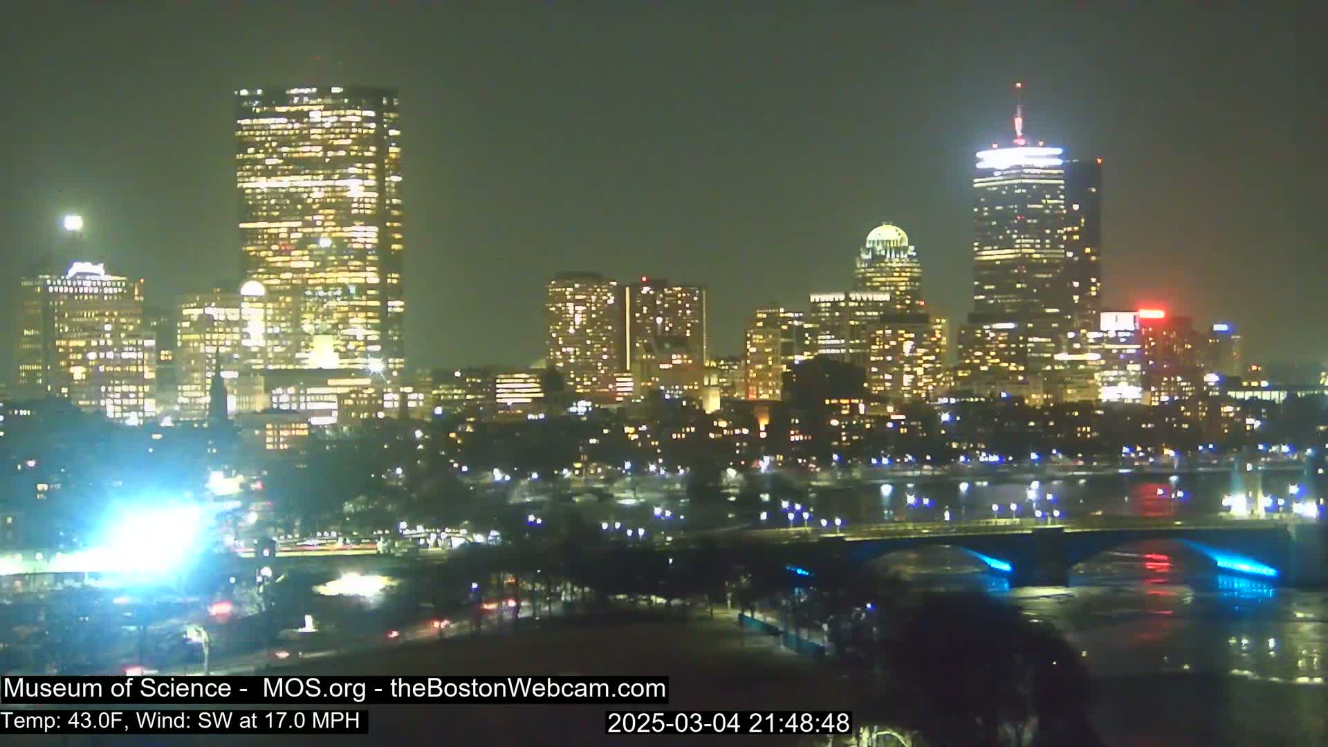 A nighttime view of a city skyline across a body of water, under a clear sky with a southwest wind at 17 mph and a temperature of 43 degrees Fahrenheit.