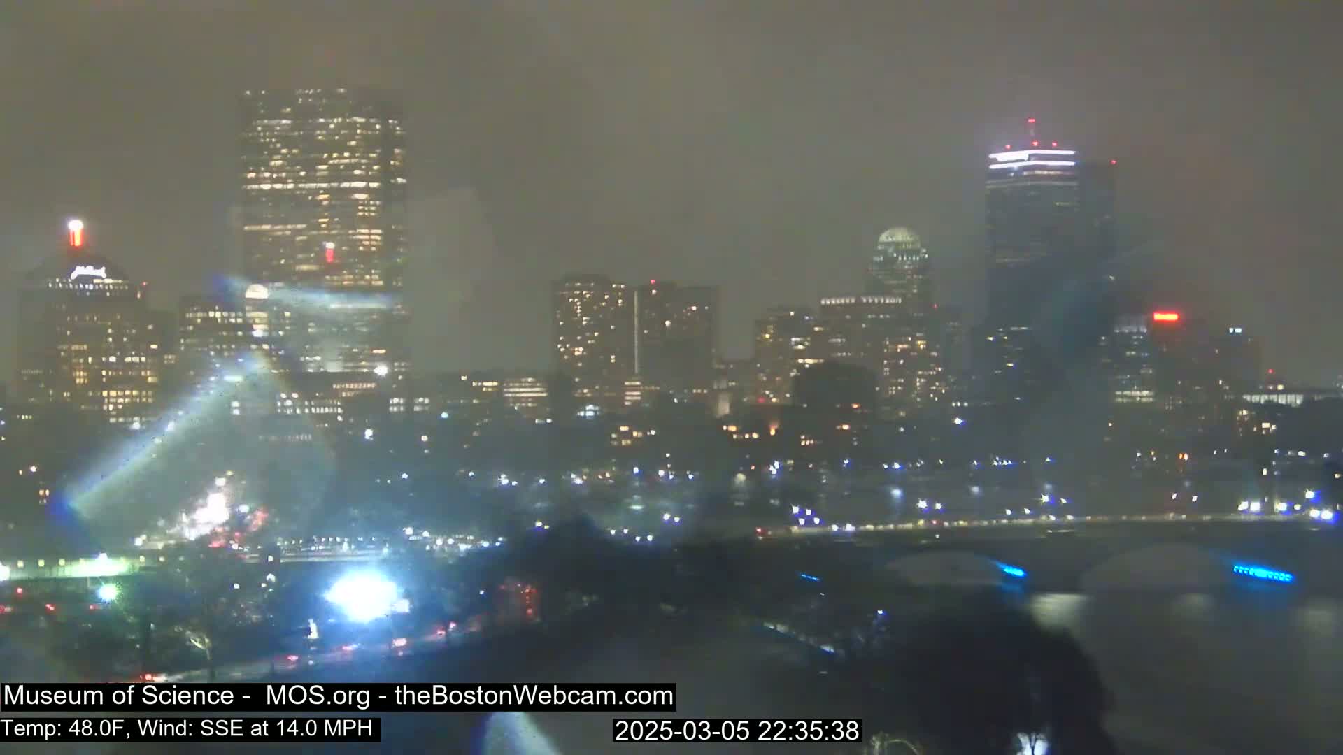 A nighttime view of a city skyline shrouded in fog or mist under windy conditions.