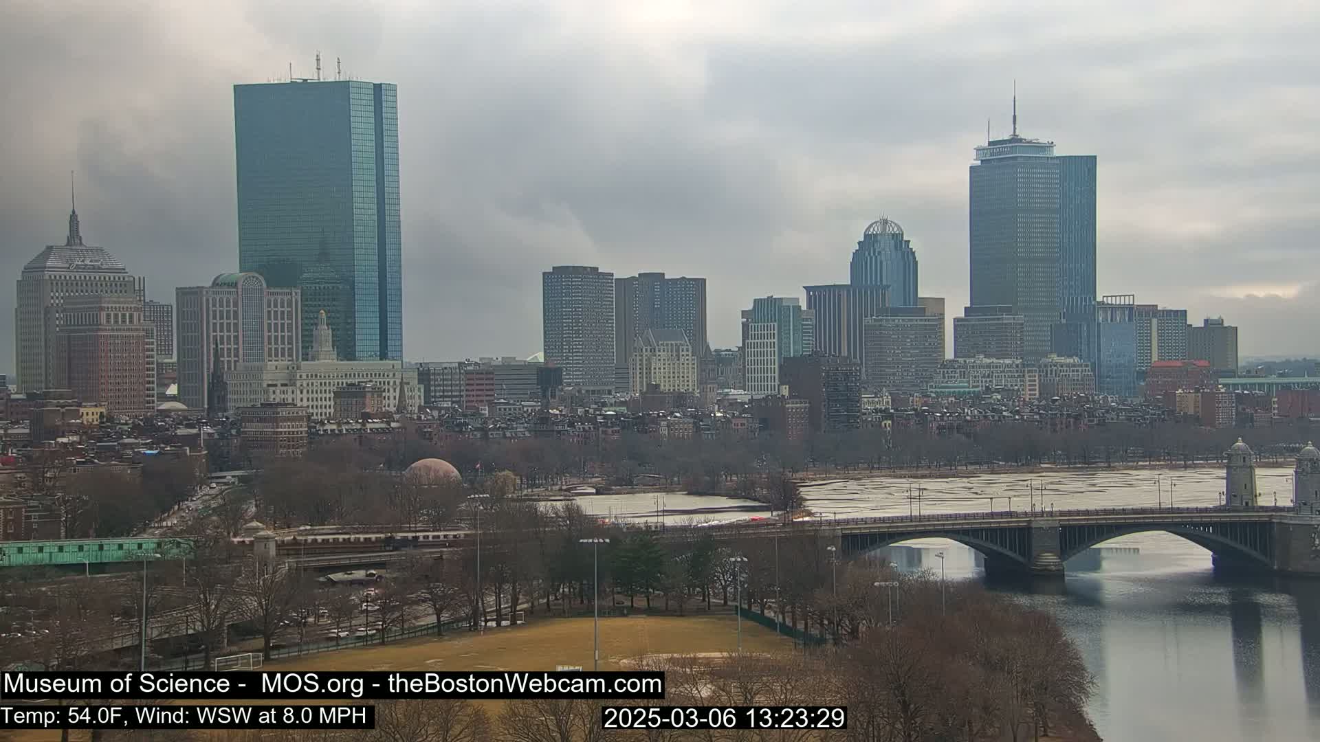 A city skyline with numerous skyscrapers and buildings is visible across a river with a bridge, under an overcast sky.