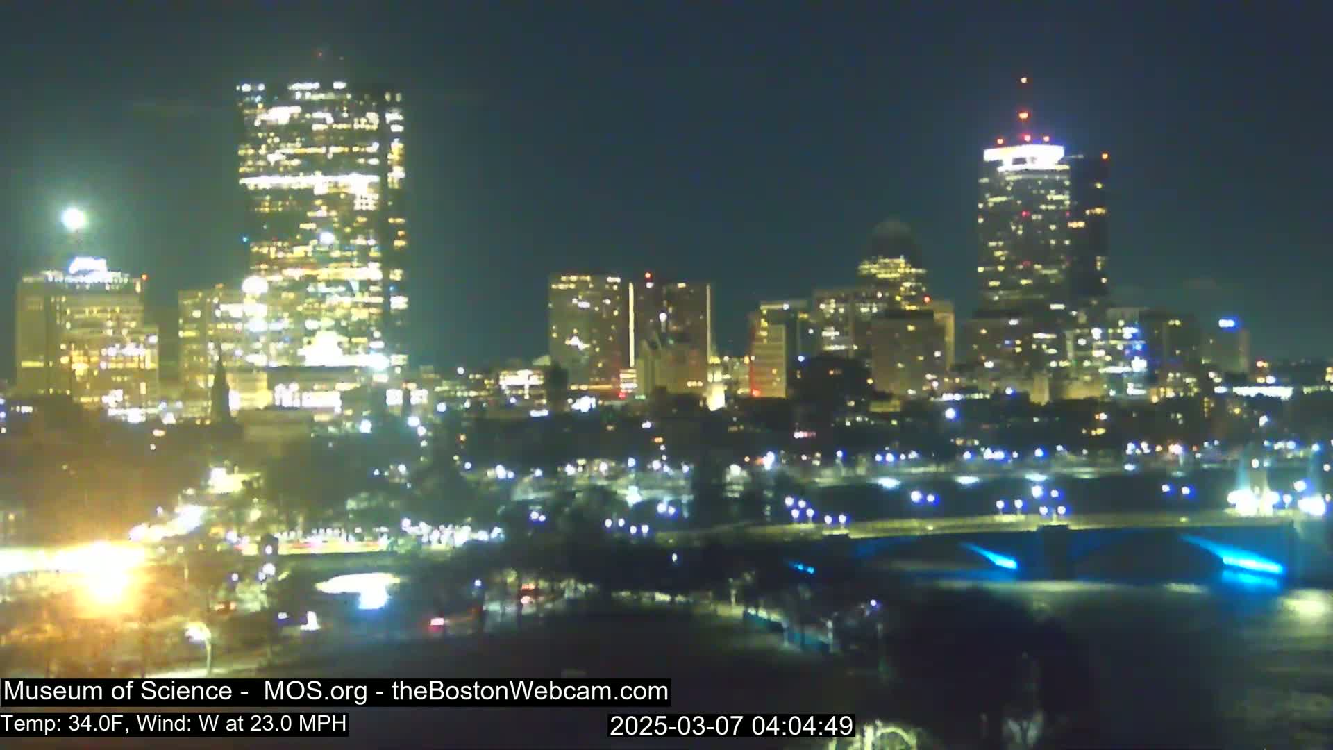 A nighttime view of a city skyline with brightly lit buildings, including a tall skyscraper, across a body of water with lights along its edge, under a clear, dark sky with a temperature of 34°F and a 23 mph west wind.