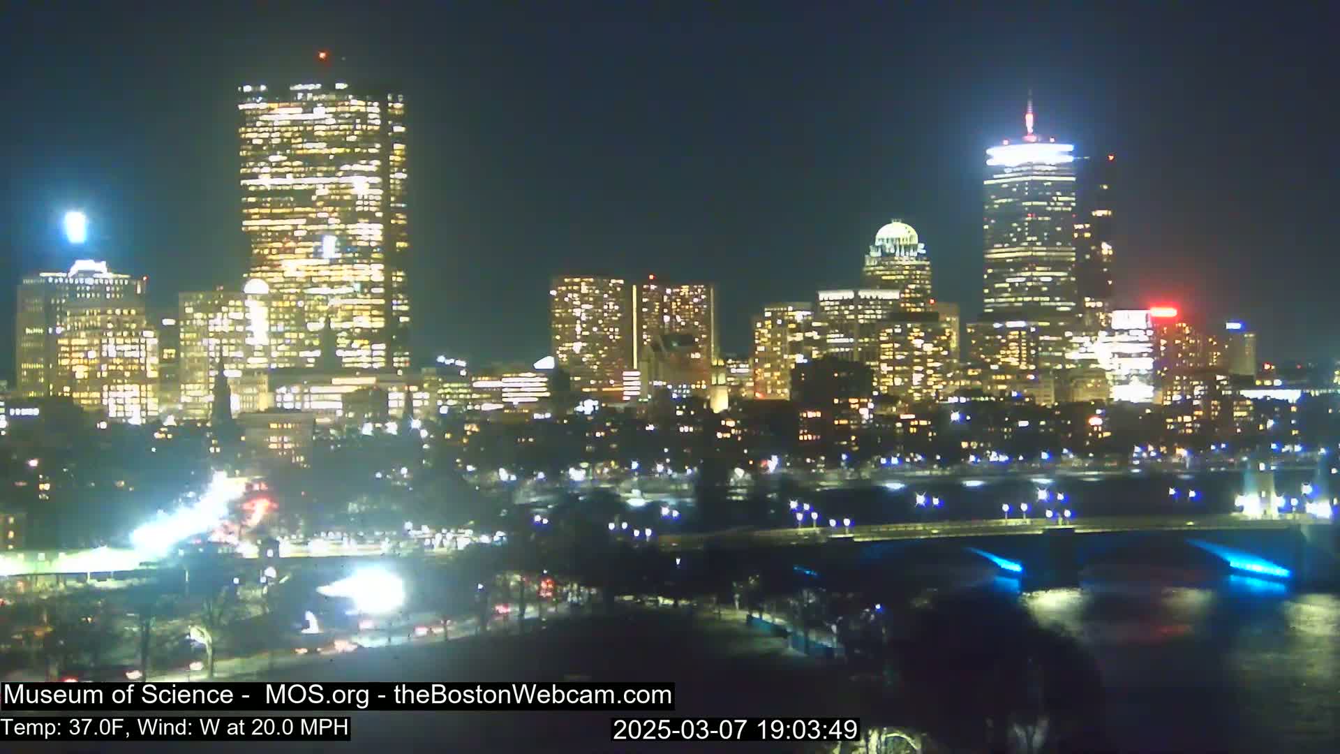 A nighttime cityscape of illuminated skyscrapers and buildings across a dark body of water, under a clear sky with a 20 mph west wind and a temperature of 37°F.