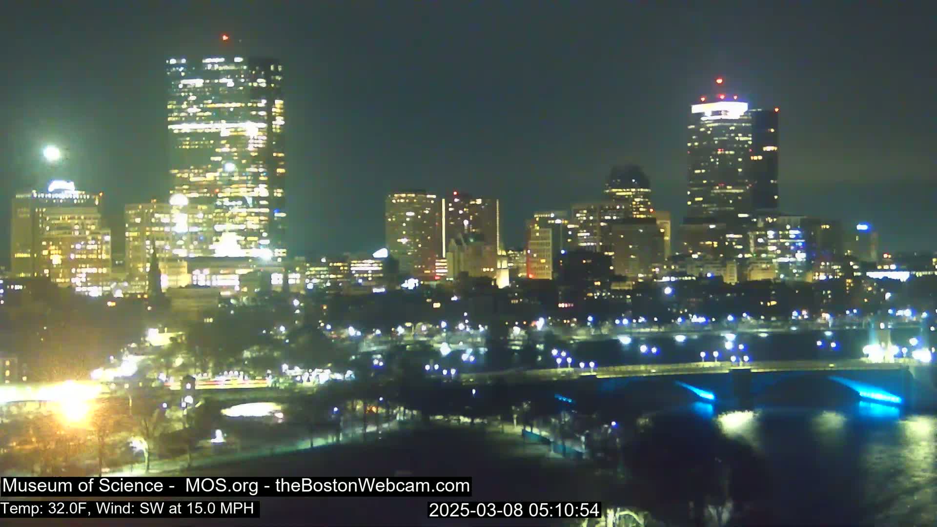 A nighttime cityscape with illuminated buildings along a river, under a partly cloudy sky with a southwest wind at 15 mph and a temperature of 32°F.