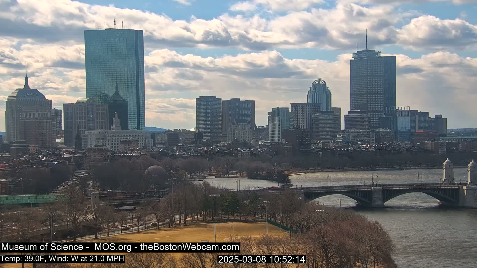 A city skyline with numerous skyscrapers is visible across a river with a bridge, under a partly cloudy sky.