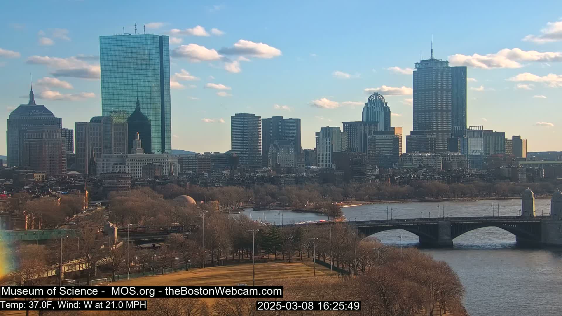 A sunny cityscape with a few clouds, featuring a river, a bridge, and many tall buildings, with bare trees lining the riverbank and a mostly clear sky.