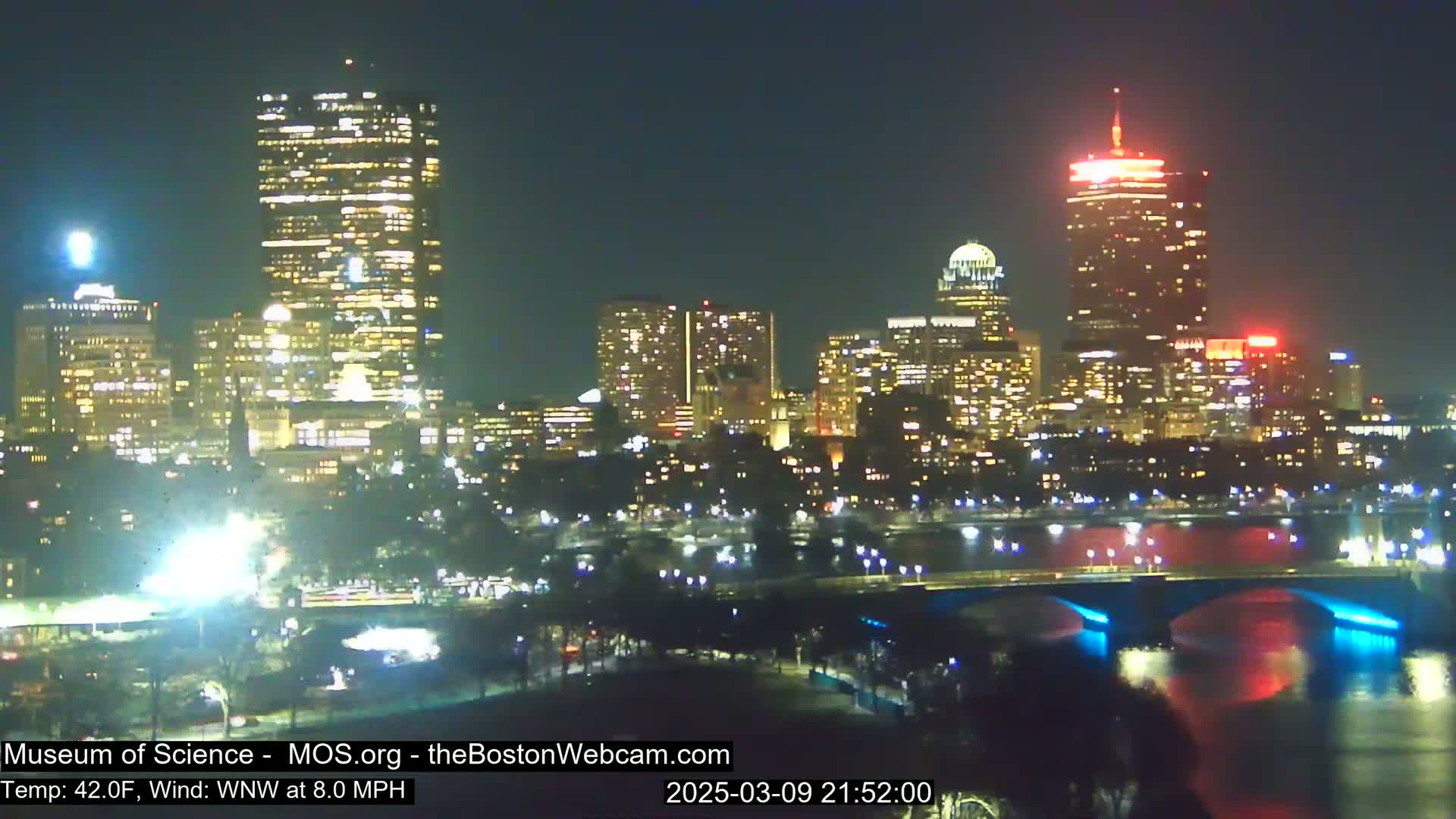 A nighttime cityscape with brightly lit skyscrapers and a bridge spanning a body of water under a clear sky, with a temperature of 42°F and a west-northwest wind at 8.0 mph.